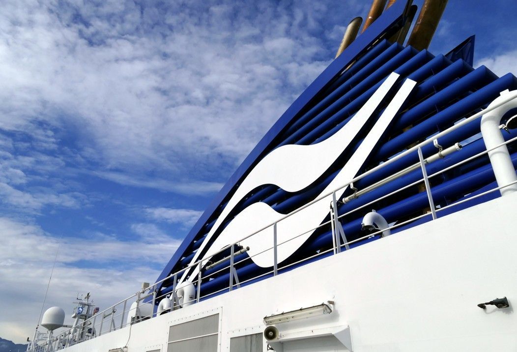 Een blauw-witte schoorsteen van een cruiseschip met een gestileerd wit logo tegen een bewolkte hemel.