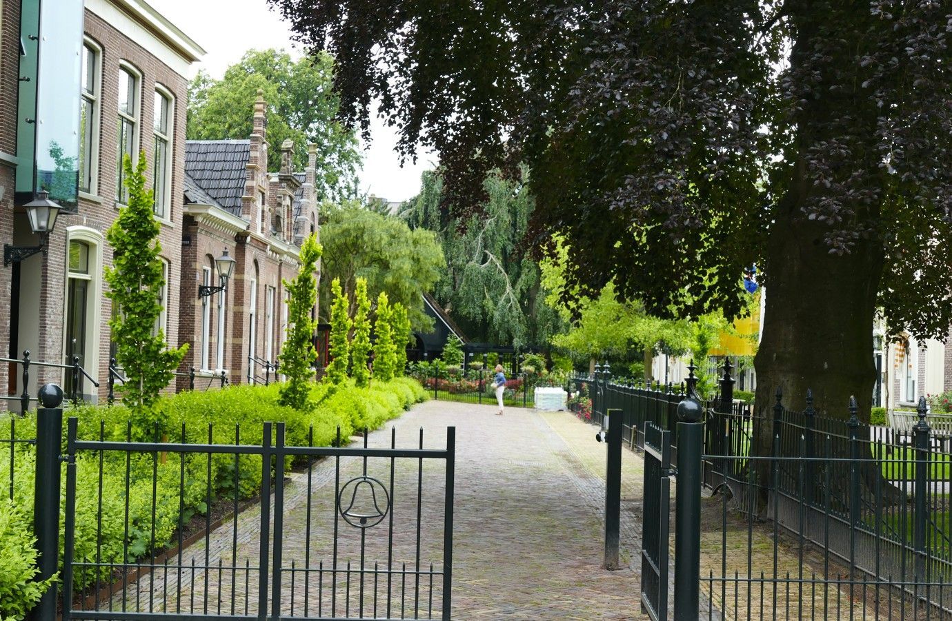 Een bakstenen pad leidt door een smeedijzeren poort, geflankeerd door hagen, bomen en gebouwen.