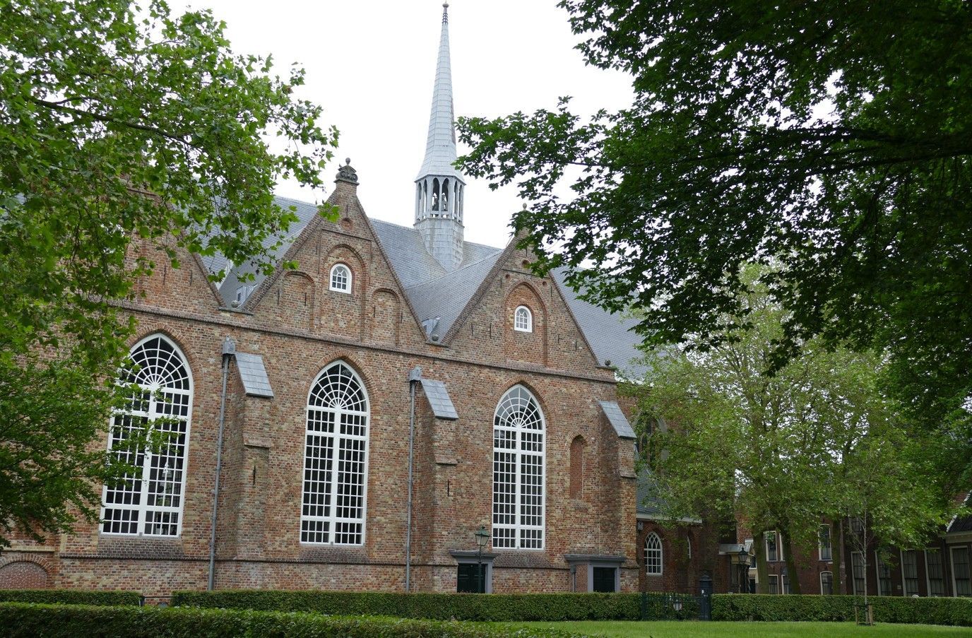 Een bakstenen gebouw met boogvensters en een hoge torenspits, zichtbaar door de bomen.