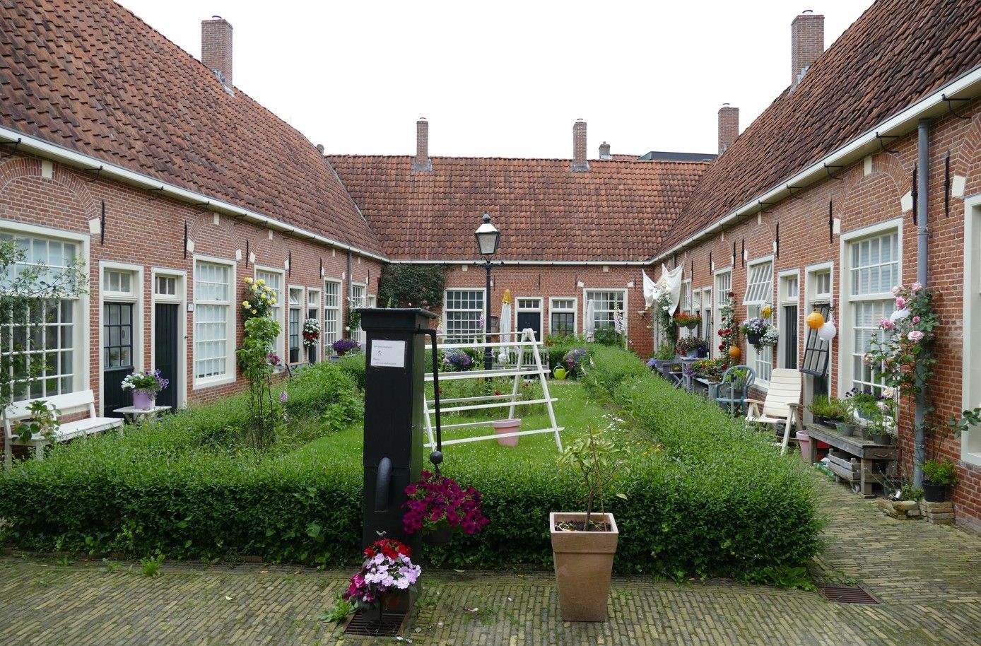 Binnenplaats met rijtjeshuizen van baksteen, rode daken en groene hagen. Een ladder staat bij een centrale lantaarnpaal.
