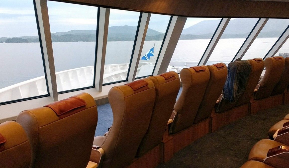 Interieur van een veerboot met bruine stoelen tegenover grote ramen met uitzicht op het water en de heuvels in de verte.