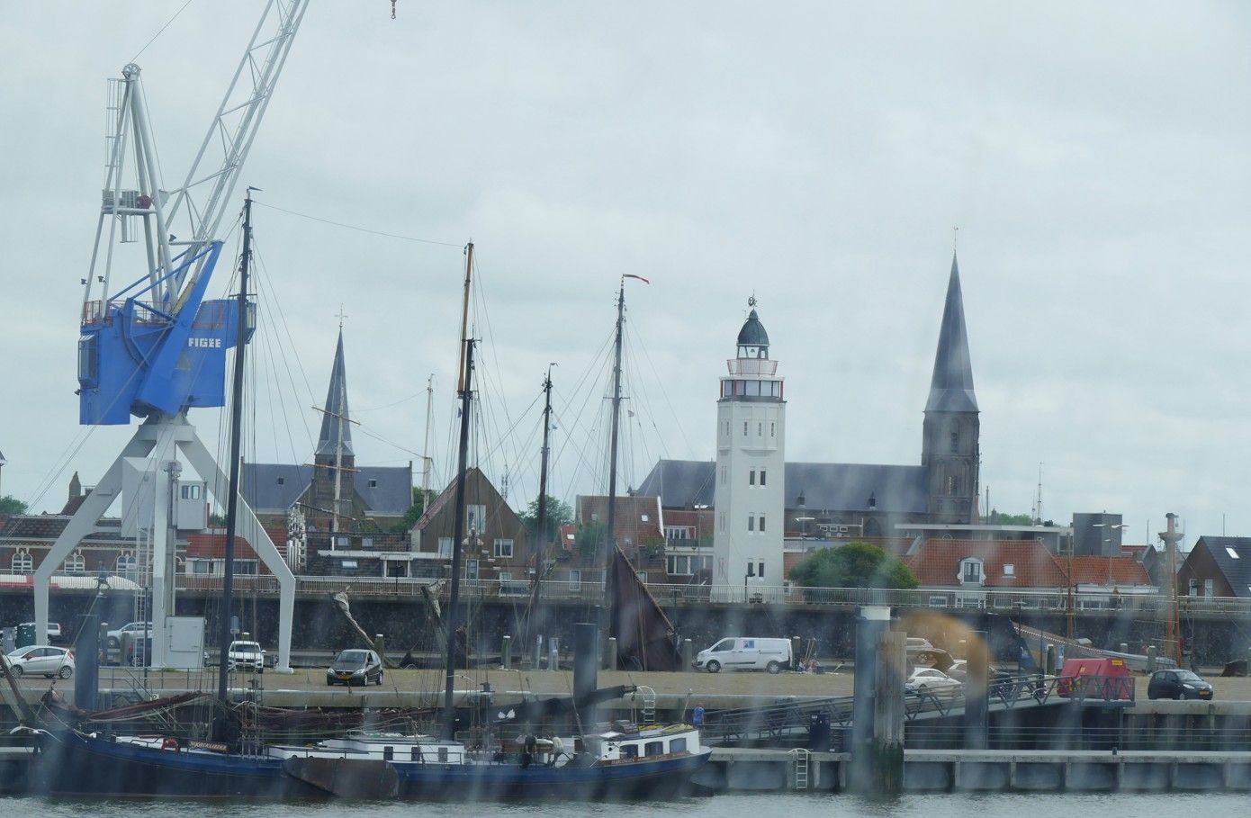 Havengezicht met een hoge blauwe kraan, masten en kerktorens onder een bewolkte hemel.