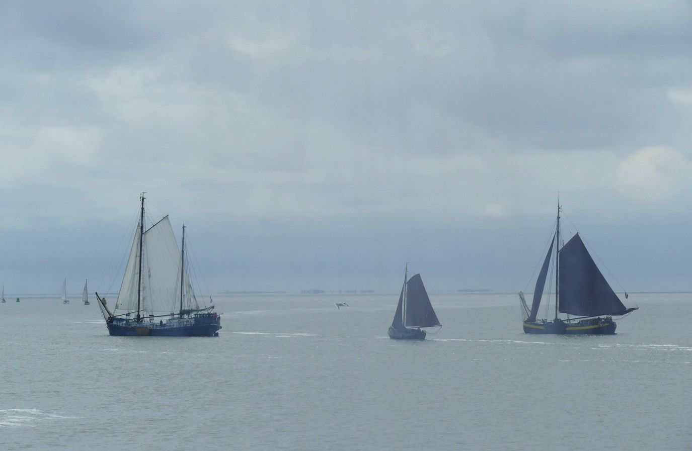 Drie zeilboten op een grijze oceaan onder een bewolkte hemel.