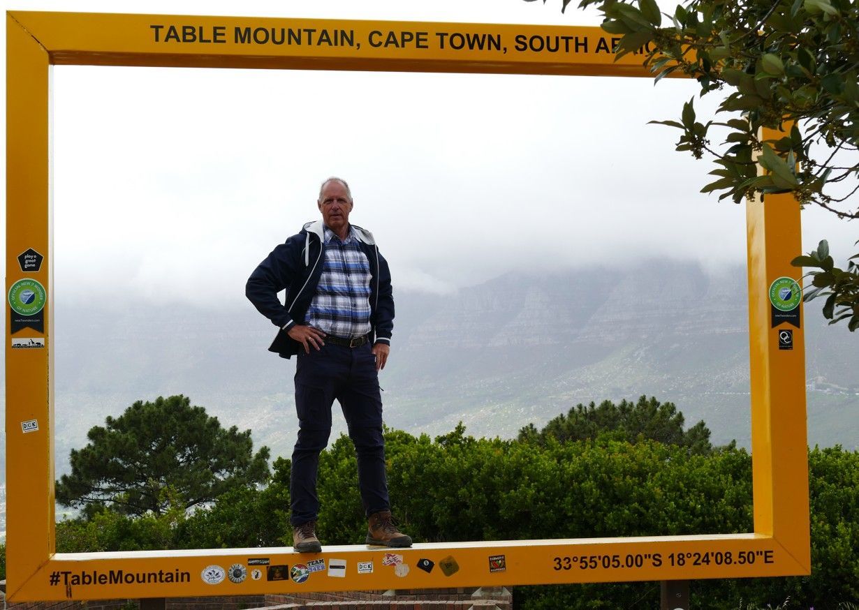 Een man poseert in een geel frame bij de Tafelberg in Kaapstad, Zuid-Afrika, met een bewolkte bergachtergrond.