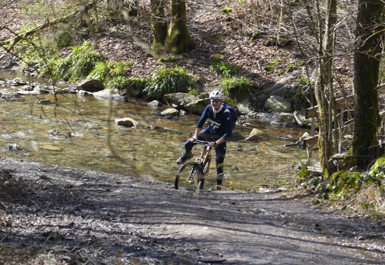 Een persoon met een helm en donkere kleding fietst op een mountainbike door een ondiepe, heldere beek in een bosrijk gebied.