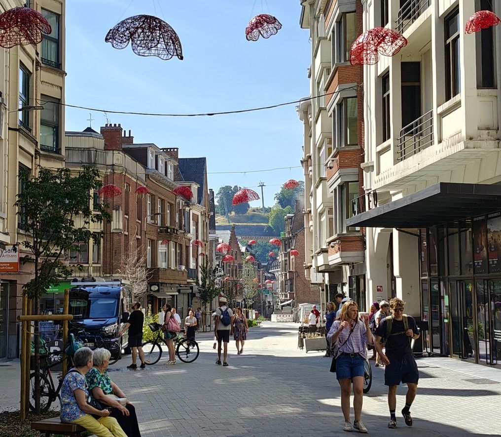 Een voetgangersstraat met mensen die wandelen, cafés en decoratieve parasols die op een zonnige dag boven het hoofd hangen.