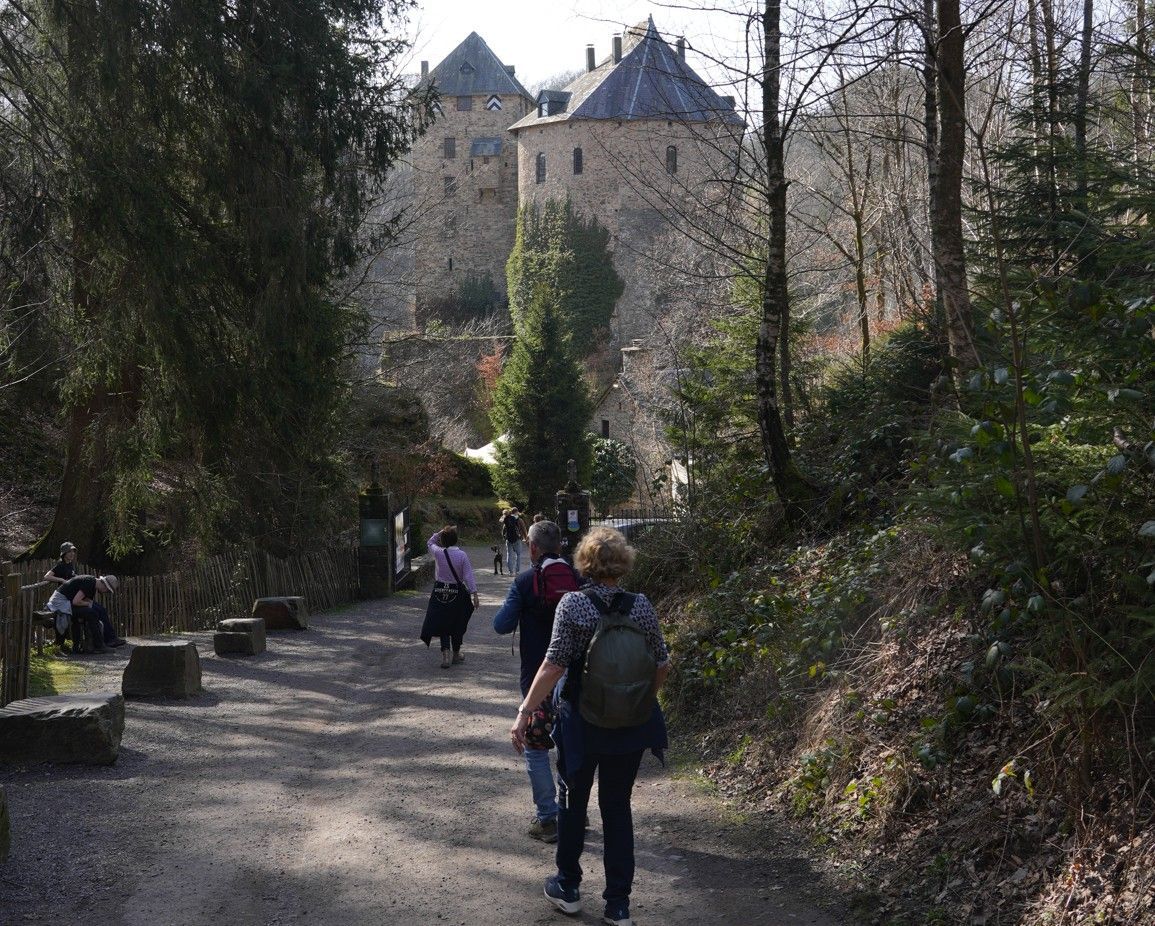 Op een zonnige dag slingert een grindpad door een bosrijk gebied naar een historisch stenen kasteel met meerdere torens.