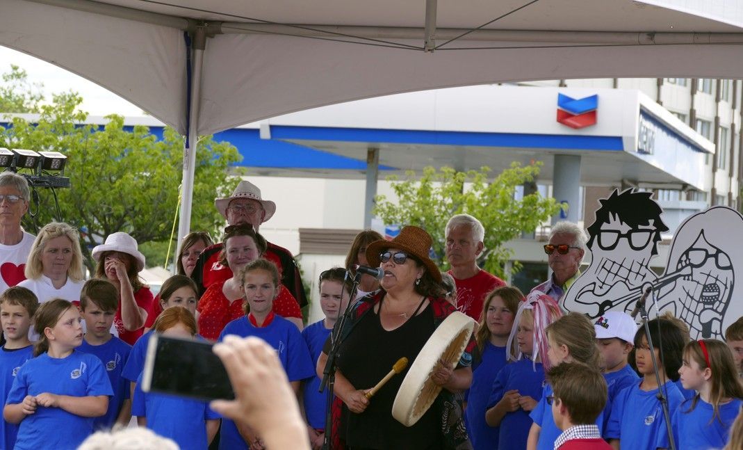 Vrouw speelt drum met kinderen in blauwe shirts; Chevron-tankstation op de achtergrond.