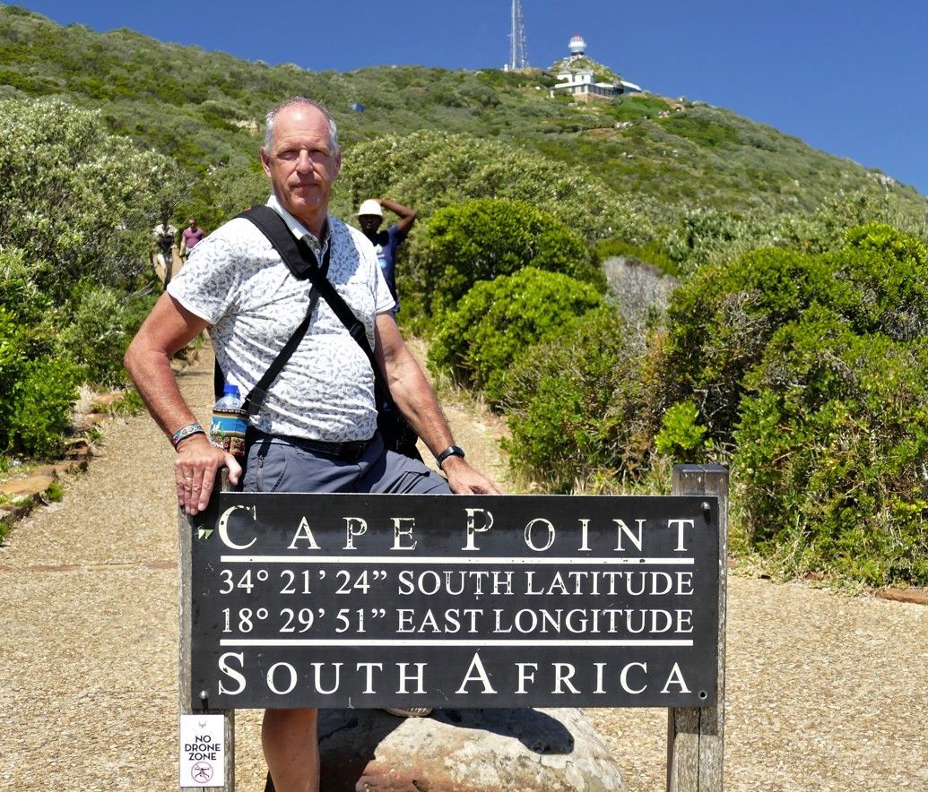 Een man bij een bord bij Kaap Punt, Zuid-Afrika. Het bord toont de breedte- en lengtegraad. Op de achtergrond zijn een heuvel en een toren te zien.