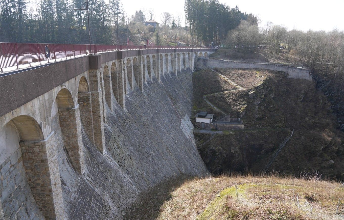 Een stenen dam met een reeks boogvormige steunberen, met een rode metalen reling langs de bovenkant.