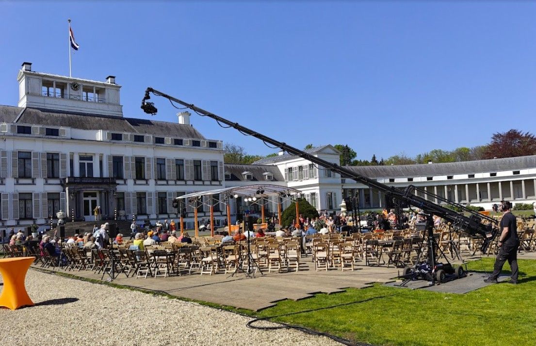 Een lange camerakraan filmt een evenement bij een groot wit gebouw met een vlag; mensen zitten aan tafels.