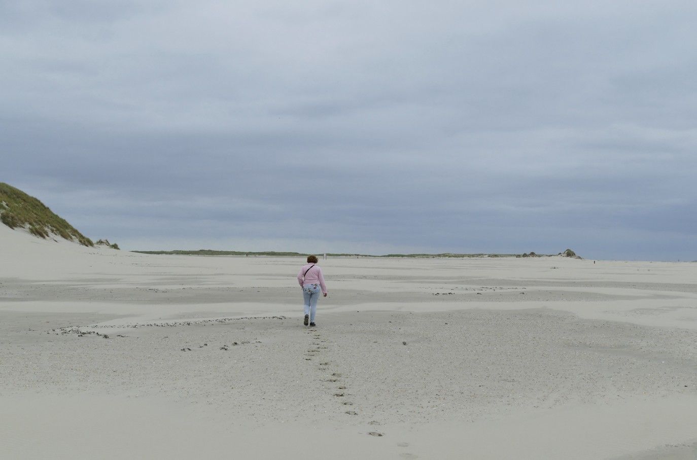Een persoon loopt over een zandstrand onder een bewolkte hemel en laat voetsporen achter.