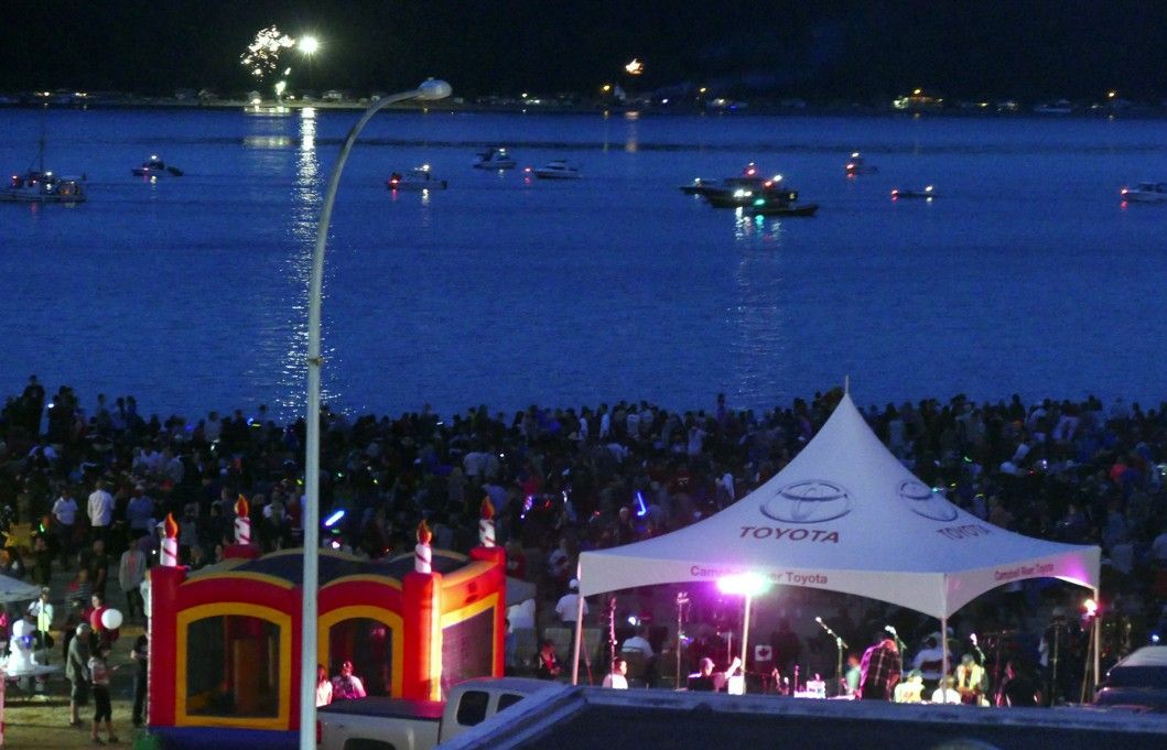 Menigte kijkt naar vuurwerk boven het water; podium met een band onder een tent; springkussen.