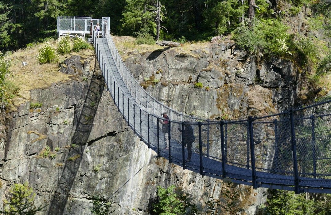 Een hangbrug overspant een rotsachtige klif. Mensen lopen over de houten loopbrug, met bossen op de achtergrond.