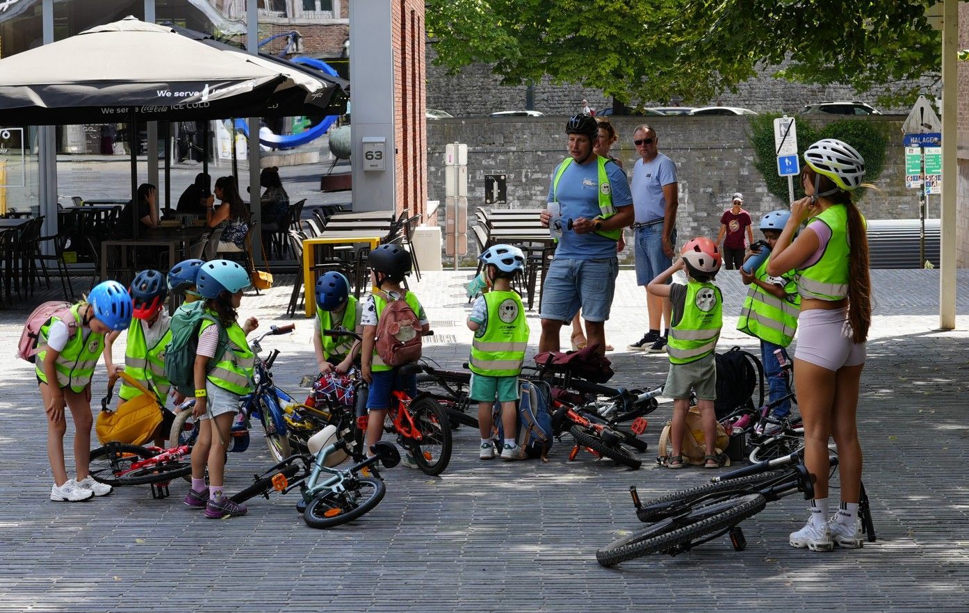 Fietsers in kleurrijke kleding rusten uit op een natte straat, vlakbij toeschouwers en hun fietsen, onder een bewolkte hemel.