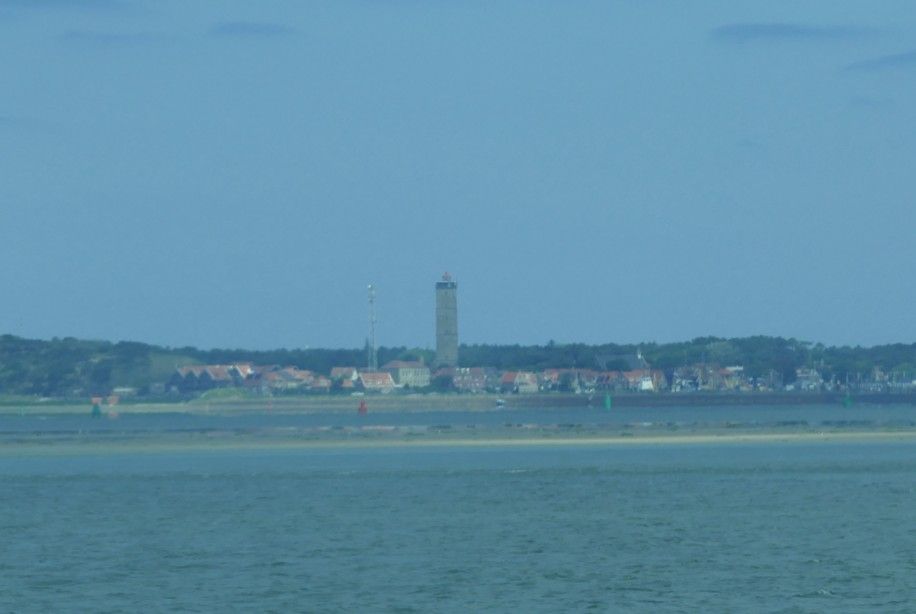 Een kustplaats met een hoge toren in het centrum, gezien vanaf de overkant van het water onder een blauwe hemel.