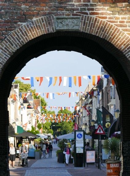Uitzicht door een bakstenen boog op een voetgangersstraat met winkels, vlaggen en mensen.