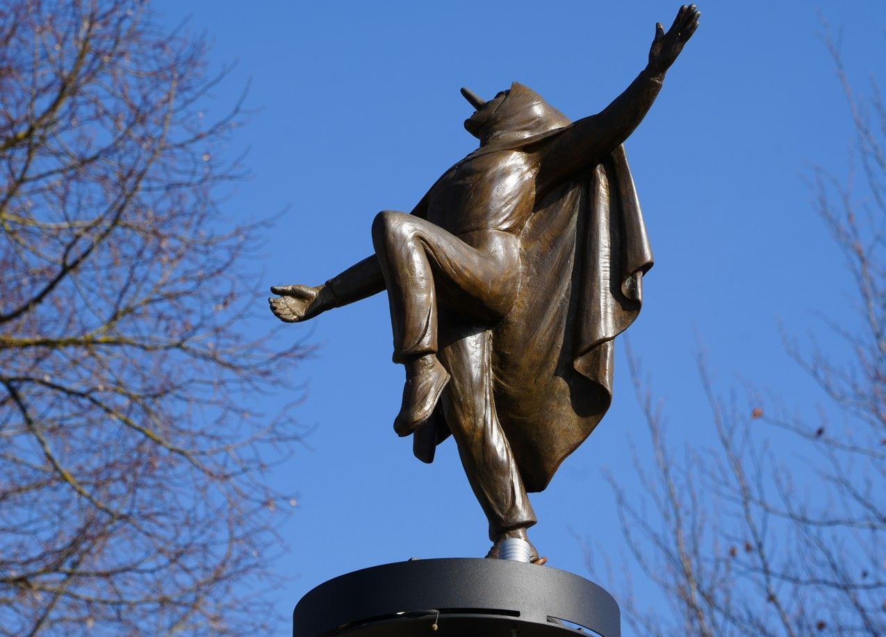 Een bronzen beeld van een persoon in een cape, afgebeeld midden in een sprong met uitgestrekte armen. Stavelot