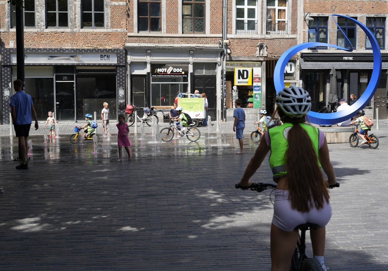 Fietsers op een stadsplein met een blauwe sculptuur en fonteinsproeiers vlakbij winkels.