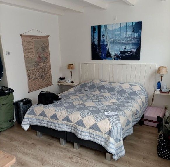 Slaapkamer met bed, dekbed, kaart, kunstwerk en bagage. Wit hoofdeinde, beige muren, houten vloer.
