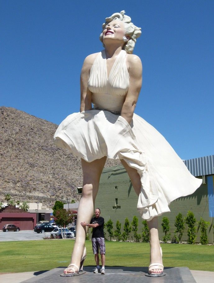 Groot standbeeld van Marilyn Monroe in een witte jurk, boven een man, voor een berg met gebouwen.