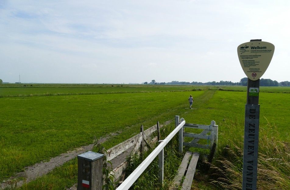 Een persoon loopt over een zandpad door een groen veld, naast een padmarkering onder een bewolkte hemel.