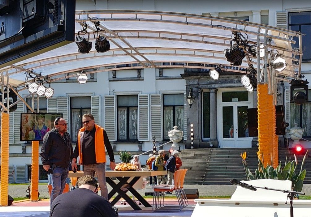 Twee mannen staan ​​op een podium met een tafel, voor een wit gebouw. ​​Ze praten met elkaar bij de productieapparatuur.
