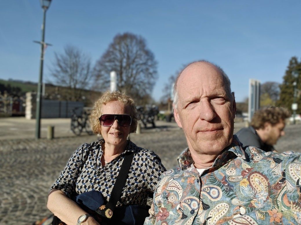 Een stel maakt een selfie in de buitenlucht op een zonnig, geplaveid plein met bomen en een lantaarnpaal op de achtergrond.