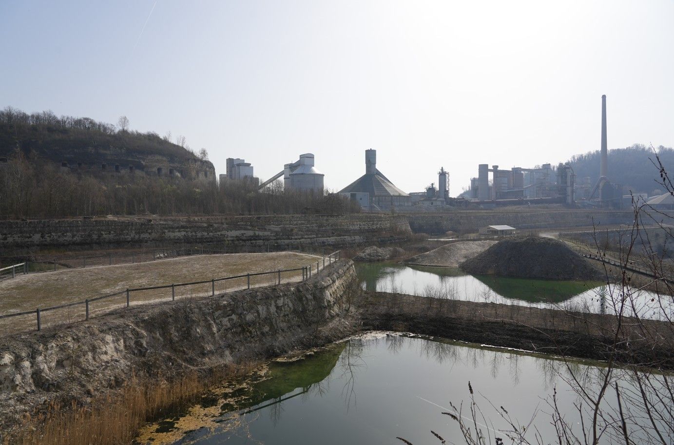 Een cementfabriekcomplex torent boven een trapsgewijs, rotsachtig steengroevelandschap uit, met  spiegelend water.