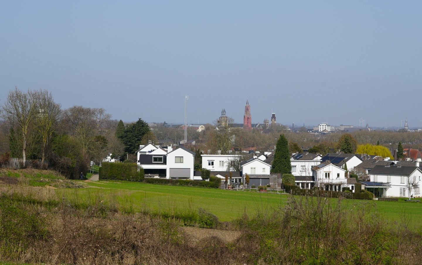 Een groen veld op de voorgrond leidt naar een groep witte huizen, met in de verte de skyline van een stad en kerktorens.