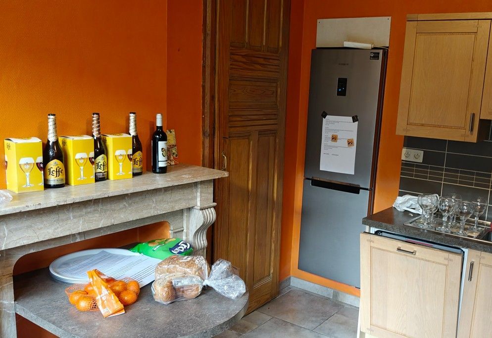 Een keuken met oranje muren, flessen en snacks op de schoorsteenmantel, een hoge koelkast en houten kasten.