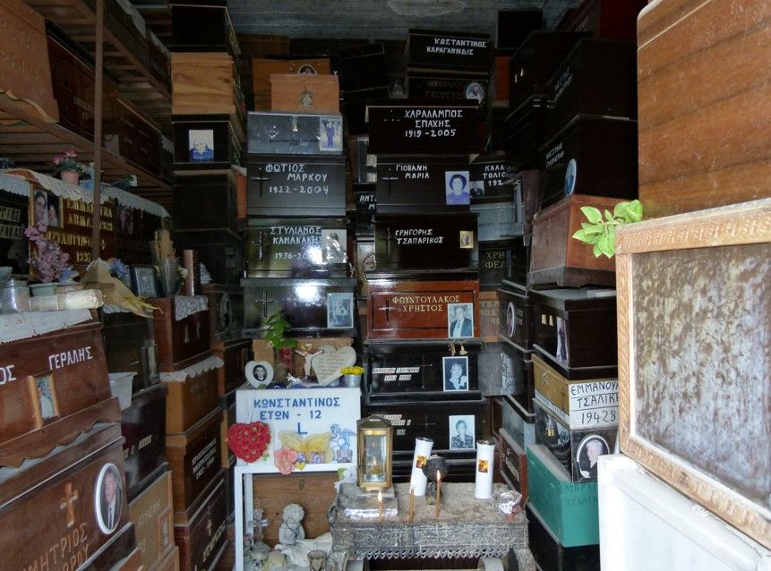 Houten doodskisten staan ​​opgestapeld in een kleine ruimte, sommige met portretten en naamplaatjes, verlicht door kaarsen en een enkele lamp.