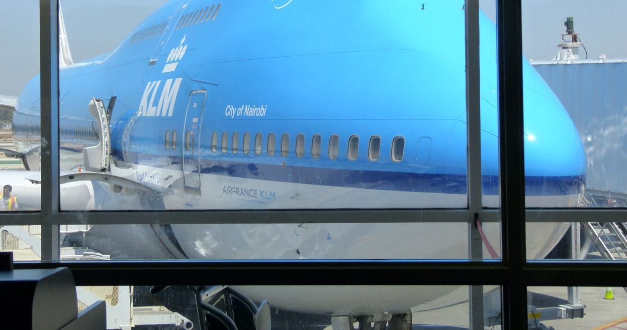 Een blauw KLM Boeing 747-vliegtuig staat geparkeerd bij een gate op de luchthaven, gezien door een raam.