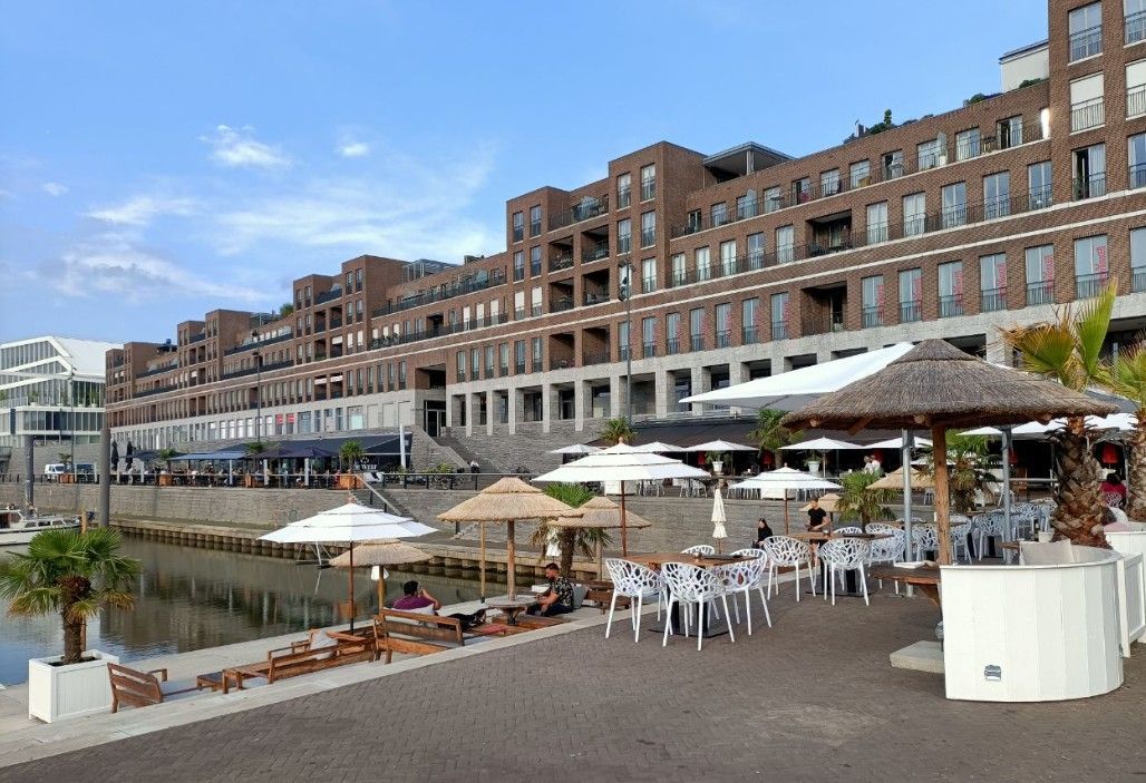 Een tafereel aan het water: een gebouw van bruine baksteen, een terras met parasols en een promenade langs het water.