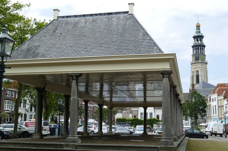 Paviljoen op een stadsplein met een kerktoren op de achtergrond en geparkeerde auto's.