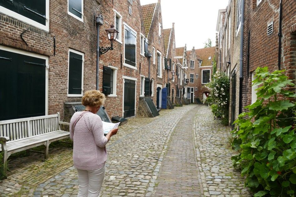 Vrouw in lichte kleding kijkt naar papier, staand op een geplaveide straat met bakstenen gebouwen.