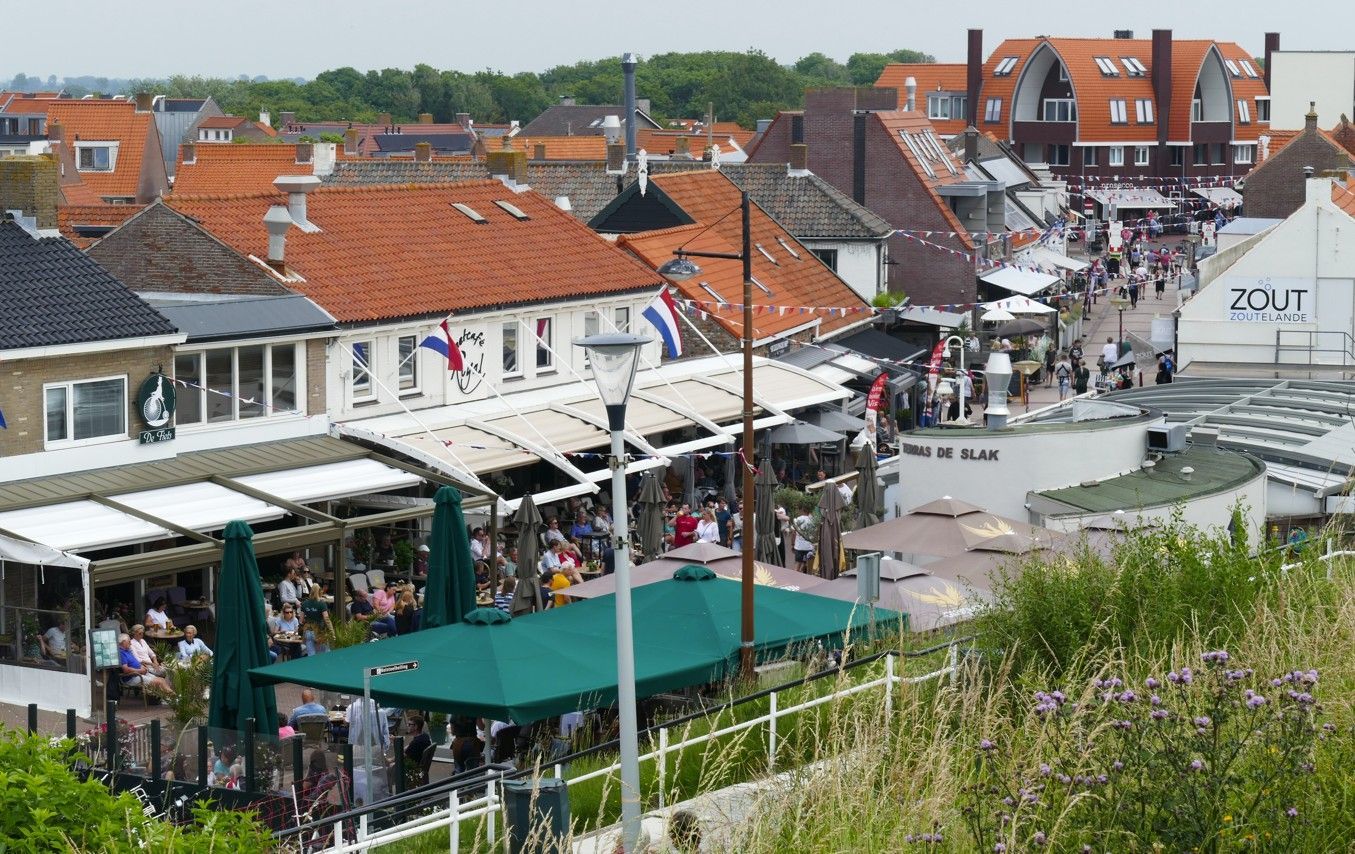 Uitzicht op een stadsplein met winkels, restaurants en mensen. Rode pannendaken, witte gebouwen en groene parasols.