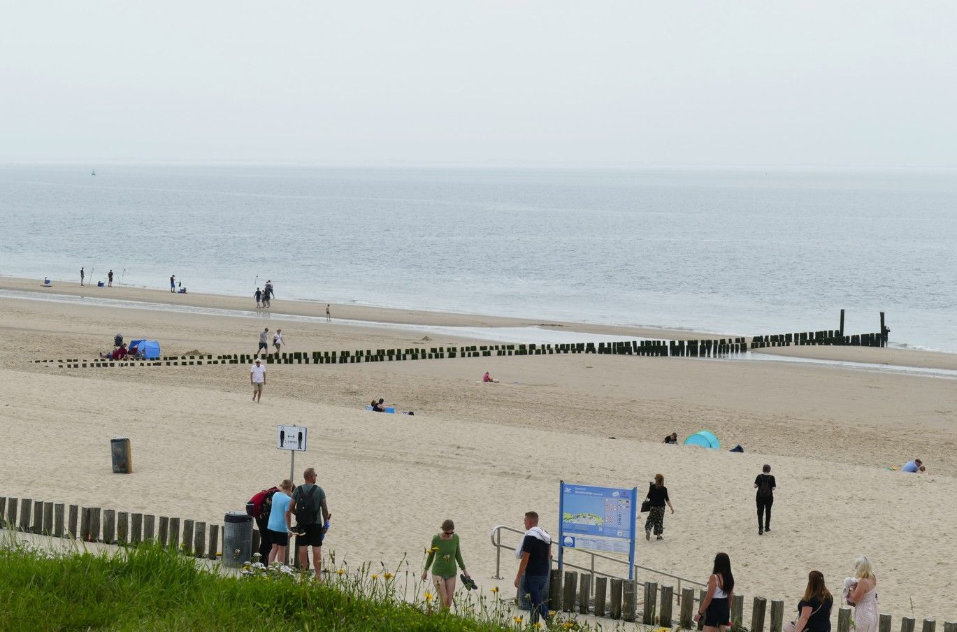 Strandtafereel met mensen op het zand, een kalme oceaan, houten golfbrekers en een bewolkte hemel.