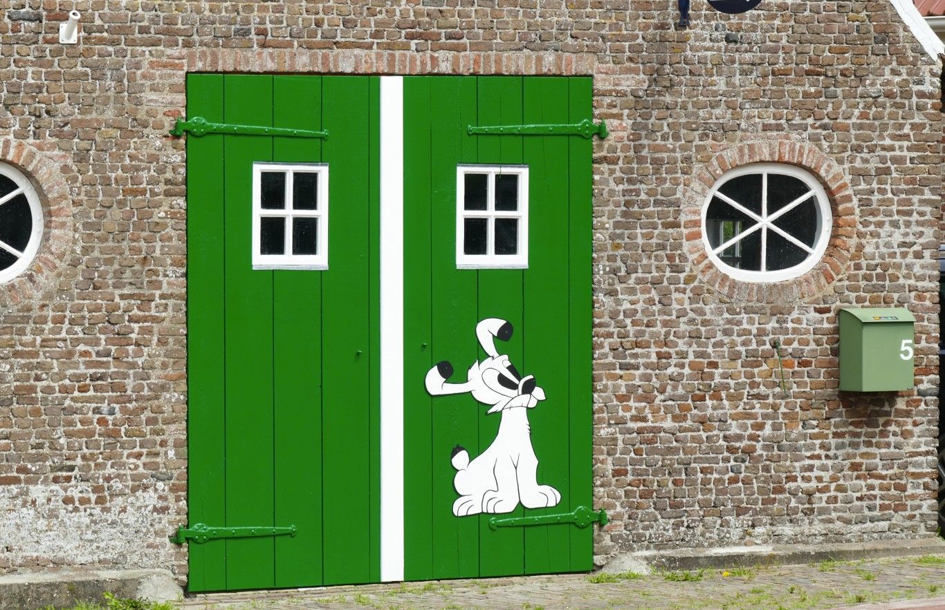 Groene houten deuren met een cartoonhondje, witte streep, kleine ramen, in een bakstenen gebouw.