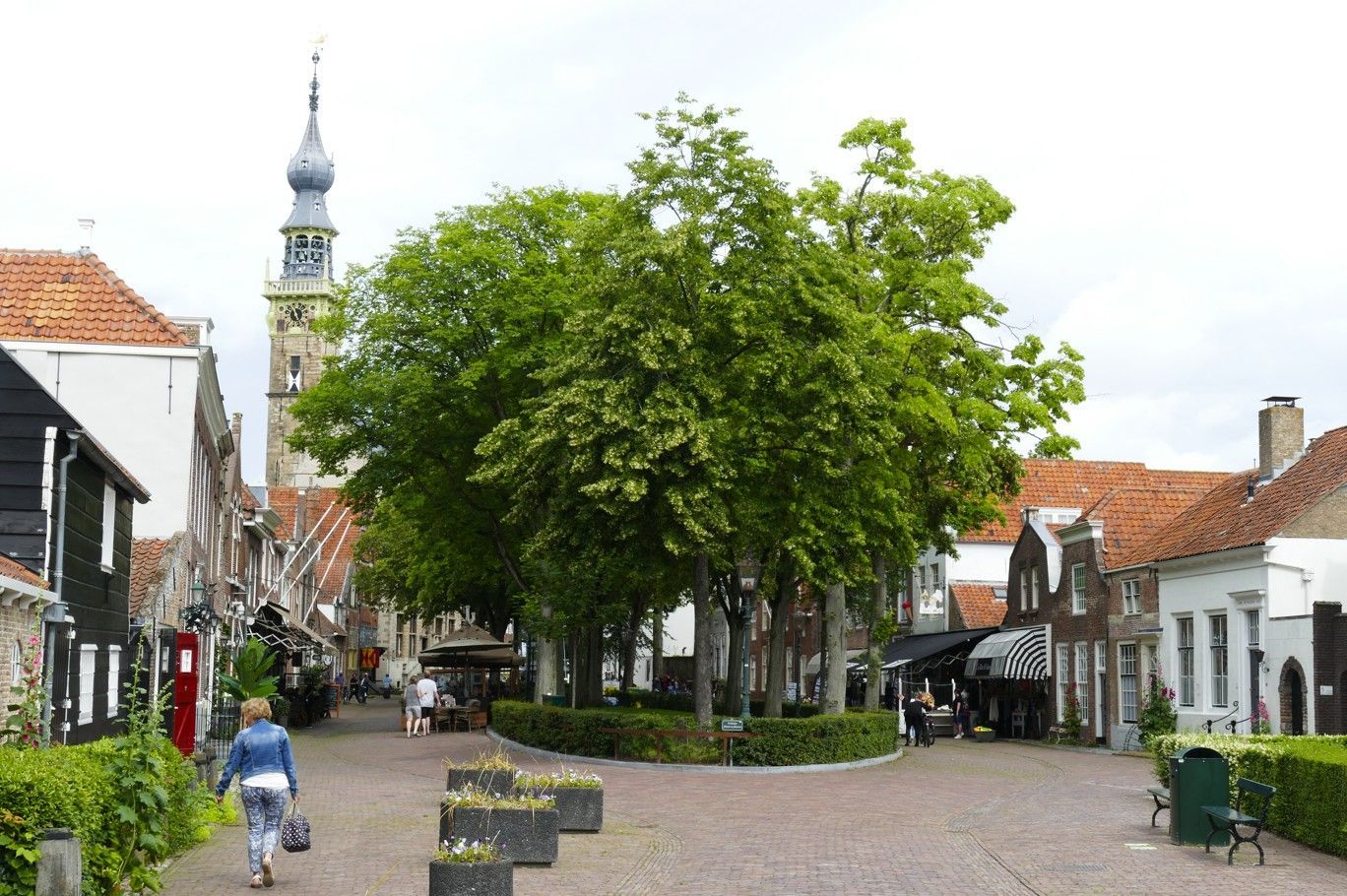 Een straat met kinderkopjes, omzoomd door gebouwen en bomen, een persoon die loopt, een kerktoren op de achtergrond.