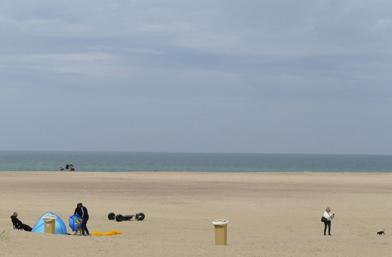 Strandscène: Mensen bij een blauwe tent, een vuilnisbak en lopend over het zand. Oceaan en lucht op de achtergrond.