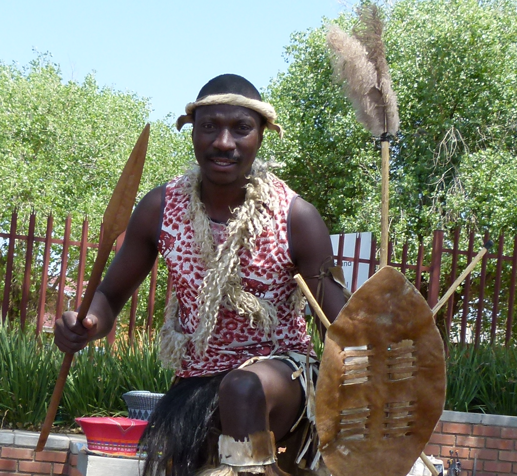 Een man in traditionele Zulu-kleding, met speer en schild in de hand, staat lachend buiten.