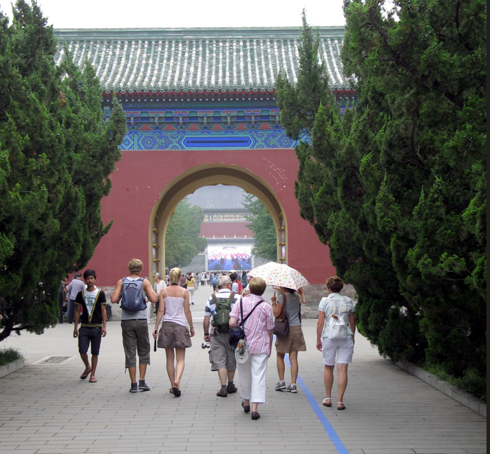 Mensen lopen richting een rode poort in een Chinees tempelcomplex, geflankeerd door groene bomen.