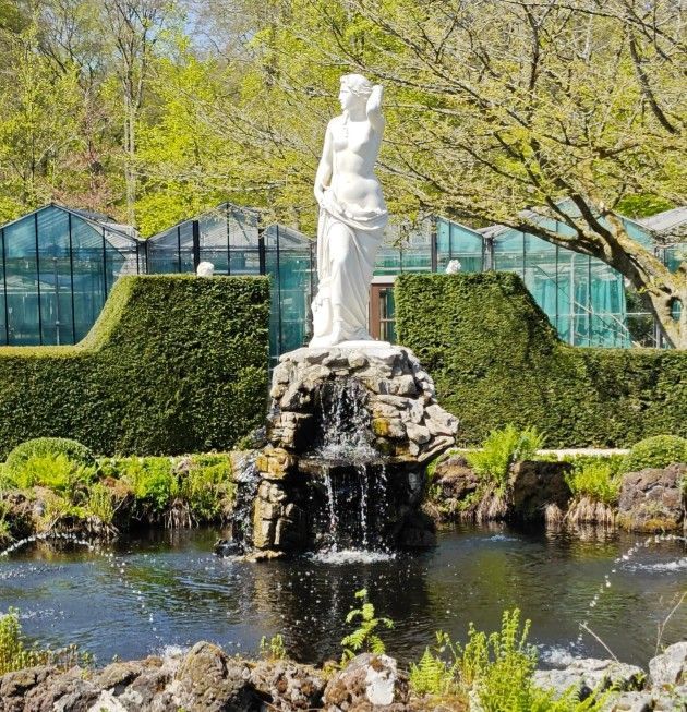 Standbeeld van een vrouw bovenop een rotsachtige waterval in een vijver, met groen en kassen op de achtergrond.
