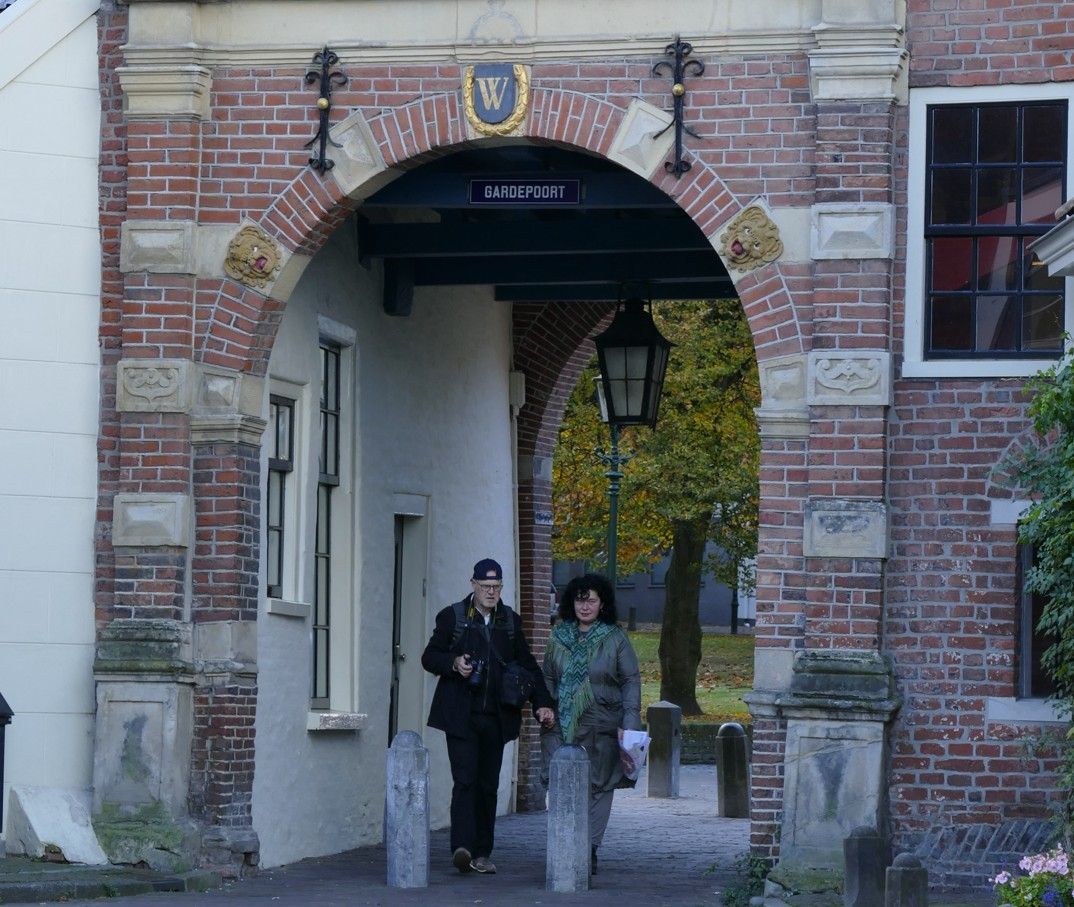 Twee mensen lopen door een bakstenen poort. Links een wit gebouw, op de achtergrond bomen.