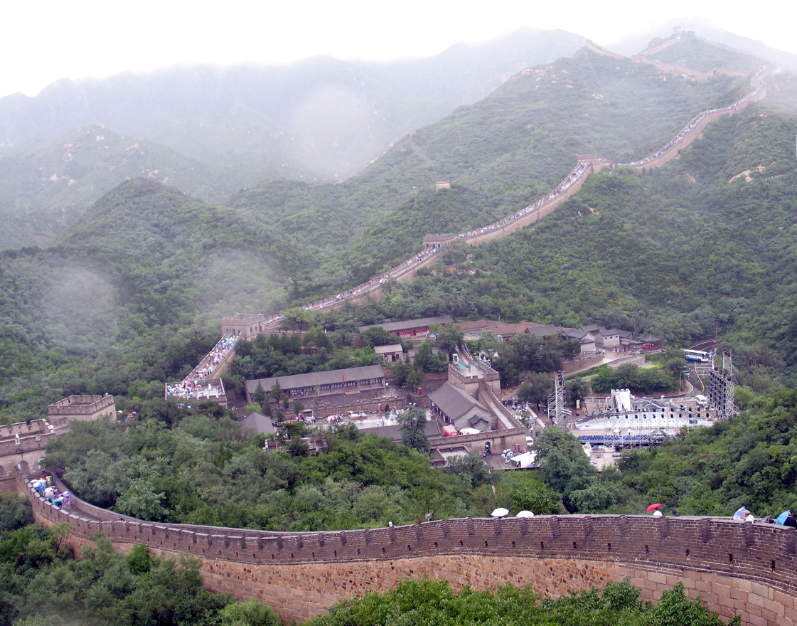 De Grote Muur van China slingert zich door een groen, bergachtig landschap, met een dorpje eronder verscholen.