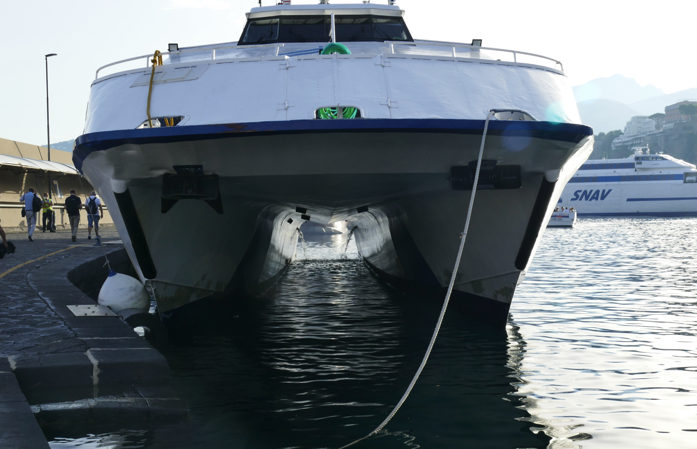Een witte catamaran met donkerblauwe accenten ligt aangemeerd aan een pier, mensen aan wal, andere boten op de achtergrond.