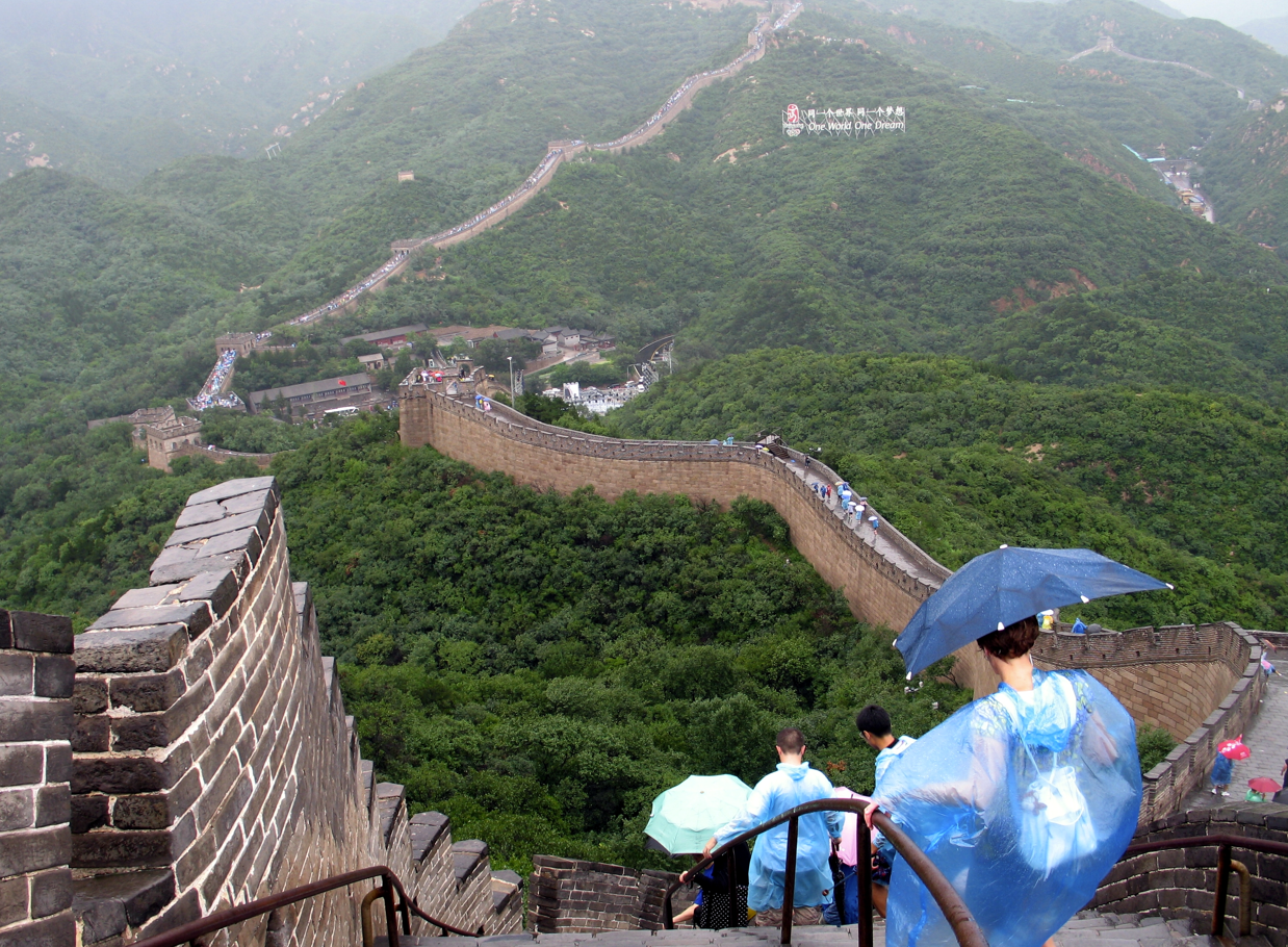 De Grote Muur van China in een groen, heuvelachtig landschap. Toeristen wandelen over de muur, sommigen met een paraplu.