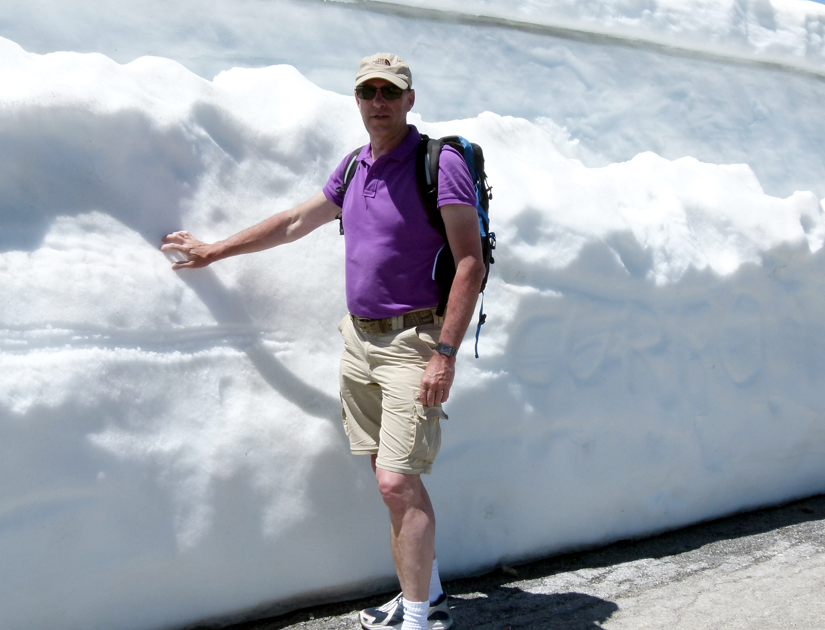 Een man in een paars shirt en kaki korte broek raakt een hoge sneeuwbank aan.
