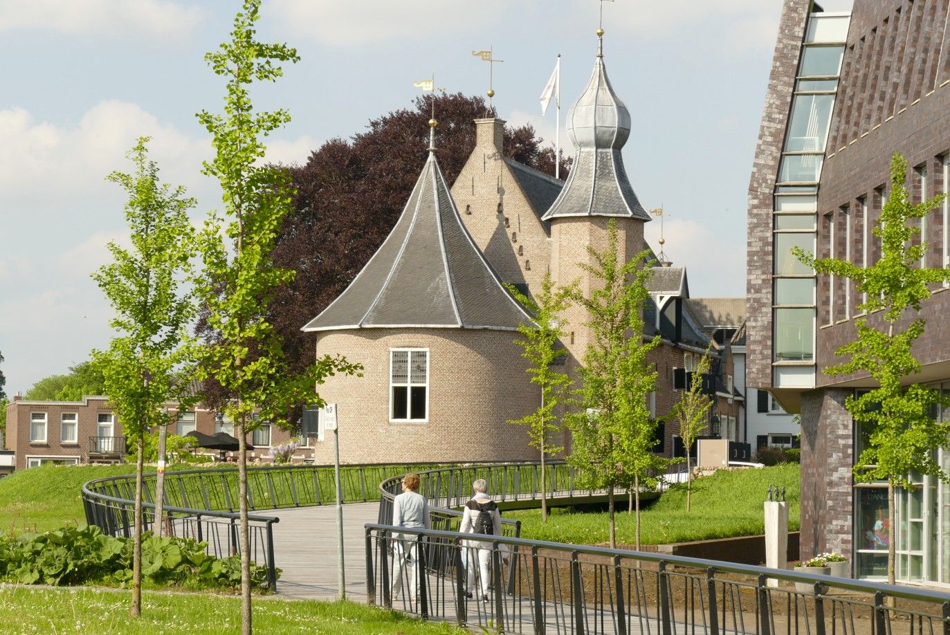 Kasteelgebouw met wandelpad, twee mensen die lopen, groene bomen en een modern gebouw.